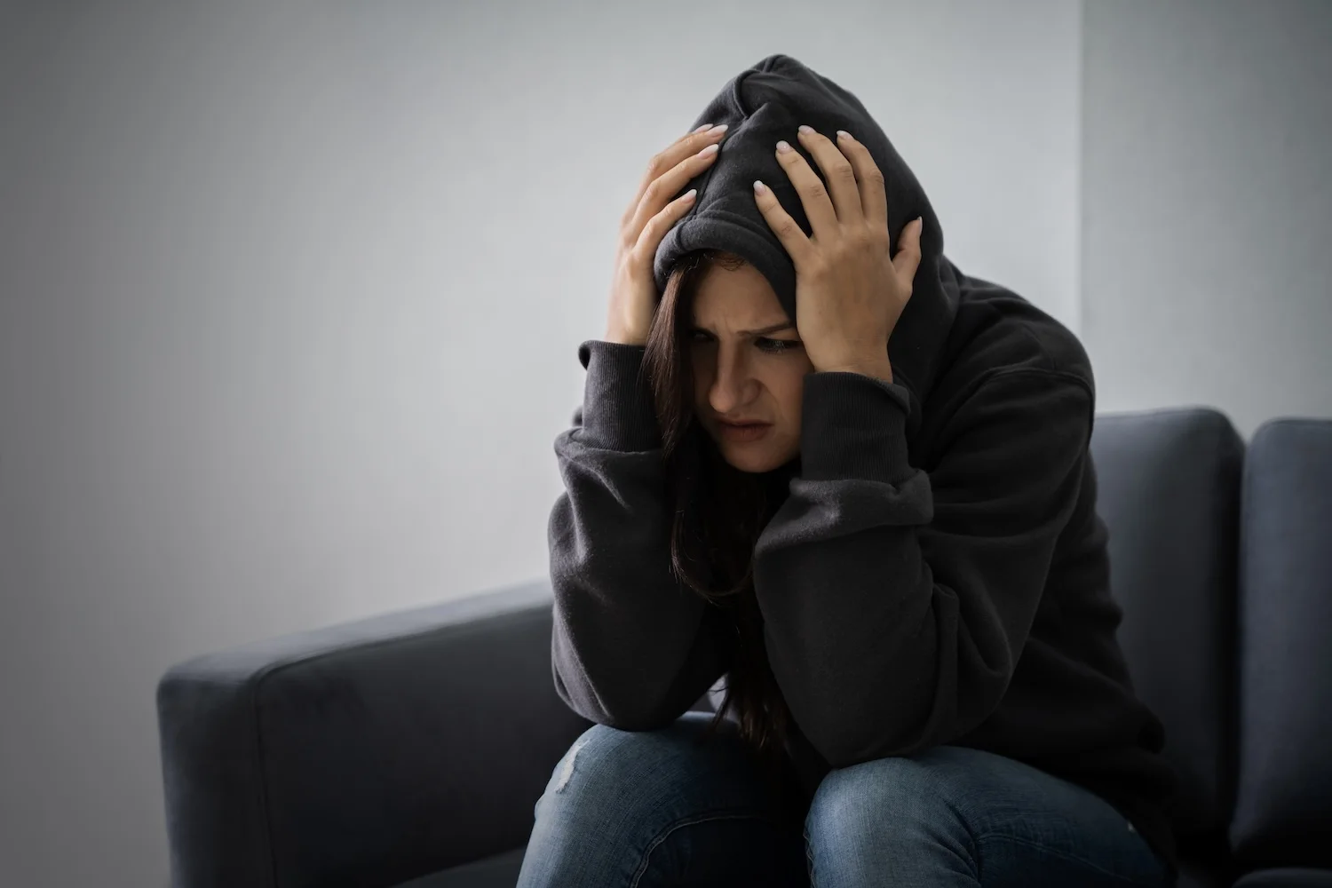 Woman sitting on a couch wearing a dark hoodie, holding her head in distress, appearing anxious or upset in a dimly lit room.