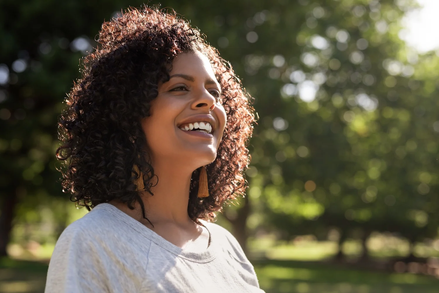 Smiling woman standing outdoors in a sunny park, wearing a light gray shirt and tassel earrings, with curly hair illuminated by natural sunlight.