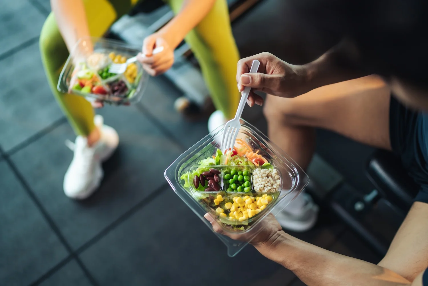 Two people in athletic clothing sitting on gym equipment and holding clear plastic meal containers filled with colorful ingredients such as lettuce, corn, peas, beans, and grains, eating with plastic forks.