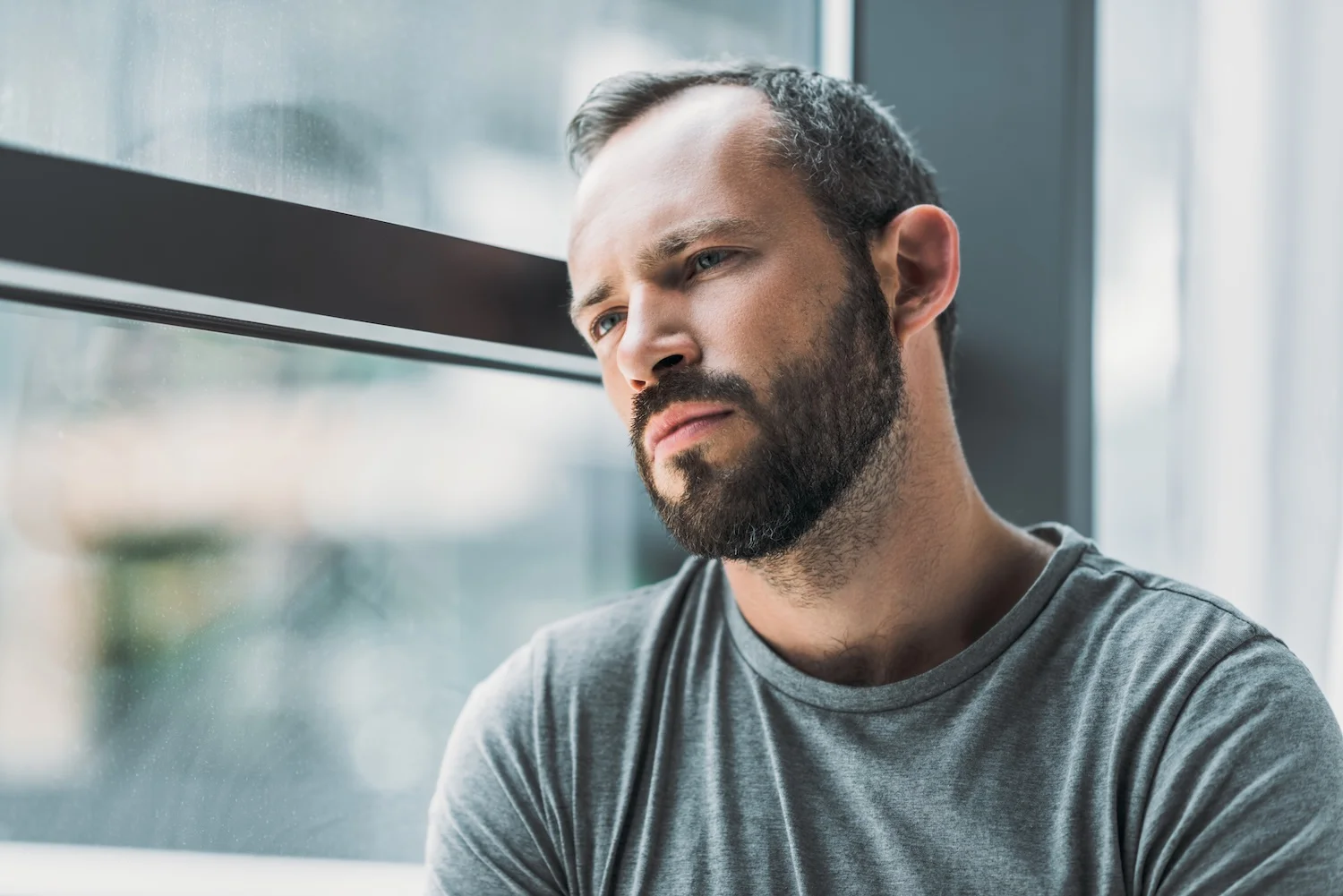 Man with a beard wearing a gray shirt looking out of a window with a thoughtful and somber expression, suggesting deep contemplation or sadness.