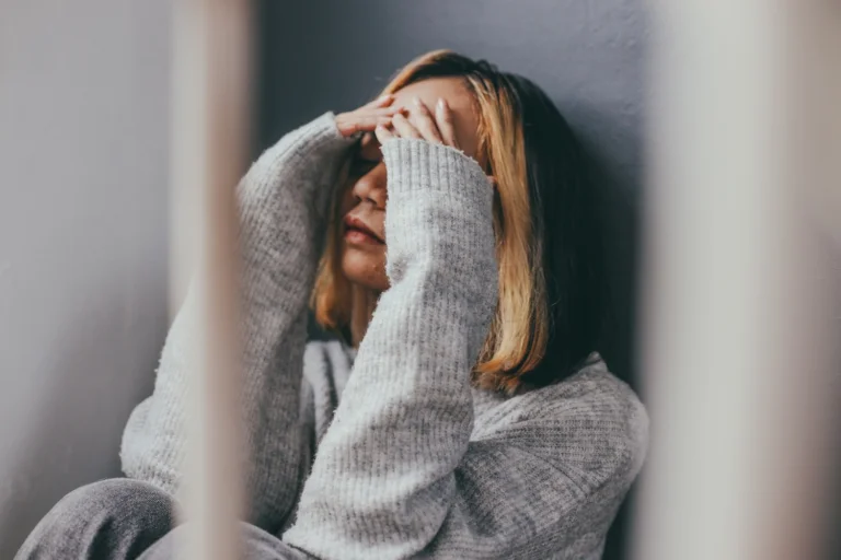 A woman sitting against a wall with her knees pulled up, covering her face with both hands. She appears distressed, wearing a gray sweater, with light framing the scene from both sides.
