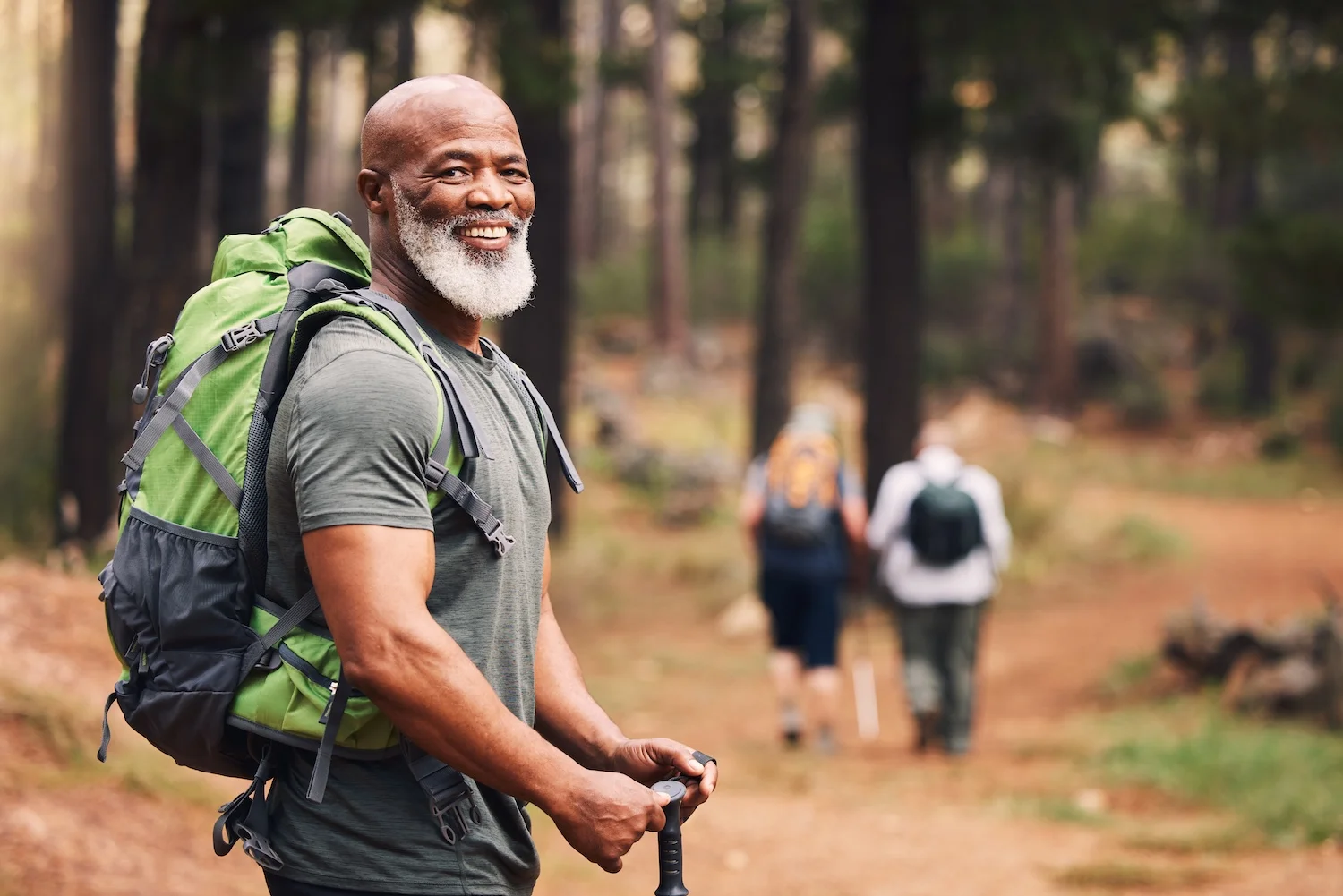 A smiling man with a gray beard stands on a forest trail, wearing a large green backpack and holding hiking poles. Two other hikers walk ahead of him in the background among tall trees.