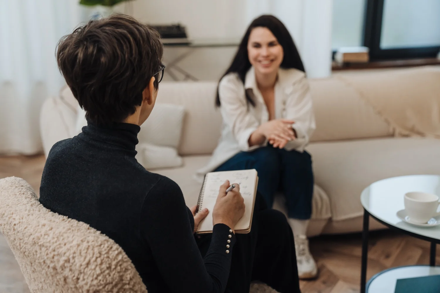 A therapist sits in a chair with a notebook, writing while facing a smiling woman who sits comfortably on a sofa. The room is bright and cozy, with soft furnishings and a cup on a small table nearby.